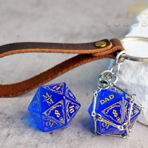 Peut inclure: Un porte-clés en cuir marron avec un anneau en métal et une cage en chaîne argentée contenant un dé à vingt faces bleu avec des chiffres dorés et le mot DAD. Un deuxième dé à vingt faces bleu avec des chiffres dorés et les lettres M M est posé.