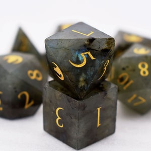 Puede incluir: Un conjunto de dados poli&eacute;dricos de varias formas, incluyendo un cubo y un d20. Los dados son de color gris oscuro con n&uacute;meros dorados. Tienen un aspecto moteado e iridiscente, con toques de azul y verde.