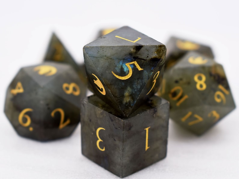Puede incluir: Un conjunto de dados poli&eacute;dricos de varias formas, incluyendo un cubo y un d20. Los dados son de color gris oscuro con n&uacute;meros dorados. Tienen un aspecto moteado e iridiscente, con toques de azul y verde.