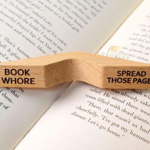 Peut inclure: Un porte-livre en bois avec deux sections. La section de gauche dit "BOOK WHORE" et la section de droite dit "SPREAD THOSE PAGES".