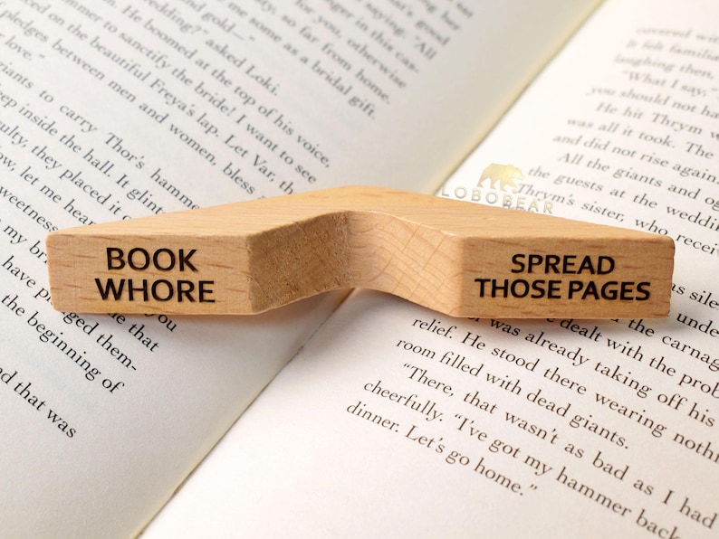 Peut inclure: Un porte-livre en bois avec deux sections. La section de gauche dit "BOOK WHORE" et la section de droite dit "SPREAD THOSE PAGES".