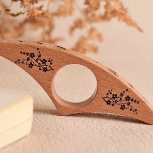 Op de afbeelding: Een houten boekenpaginahouder met een grote cirkelvormige opening en een gebogen ontwerp. De lichtbruine houder heeft zwarte bloemtakontwerpen aan elk uiteinde. De achtergrond is een neutraal beige.