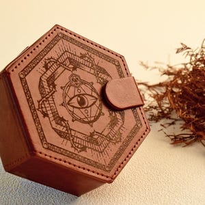 Può includere: Una scatola esagonale in pelle marrone con un design decorativo inciso con un simbolo dell'occhio. La scatola ha una chiusura a patta ed è cucita lungo i bordi. Lo sfondo include materiale vegetale secco.