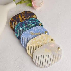 May include: Five personalised hair combs in various colours, including tortoiseshell, teal, blue, yellow, and iridescent white. Each comb has a name in gold script. The combs are arranged diagonally.