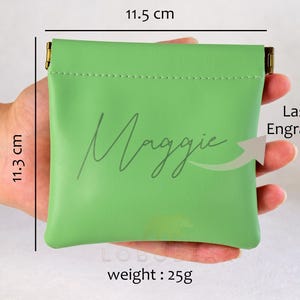 Może przedstawiać: Zielona sk&oacute;rzana kosmetyczka z zamkiem błyskawicznym. Kosmetyczka ma 11,5 cm szerokości i 11,3 cm wysokości. Na przodzie kosmetyczki wygrawerowano laserowo imię "Maggie".