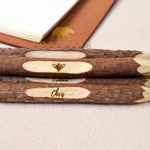 Può includere: Quattro matite di legno con finitura in corteccia naturale. Ogni matita ha un'iscrizione personalizzata e un piccolo disegno di un'ape. Le matite sono impilate l'una sull'altra.