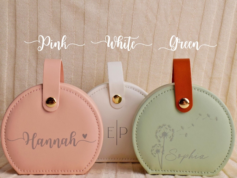 K&ouml;nnte beinhalten: Drei runde, pastellfarbene Etuis mit Griffen. Das rosa Etui tr&auml;gt den Namen "Hannah". Das wei&szlig;e Etui hat die Initialen "EP". Das gr&uuml;ne Etui hat ein Pusteblumen-Design und den Namen "Sophia". Jedes Etui hat einen goldenen Knopf.