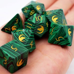 Puede incluir: Un conjunto de siete dados poli&eacute;dricos verdes y negros veteados, con n&uacute;meros dorados y s&iacute;mbolos de lobo. Los dados est&aacute;n dise&ntilde;ados para juegos de rol de mesa. Los dados se sostienen en una mano.