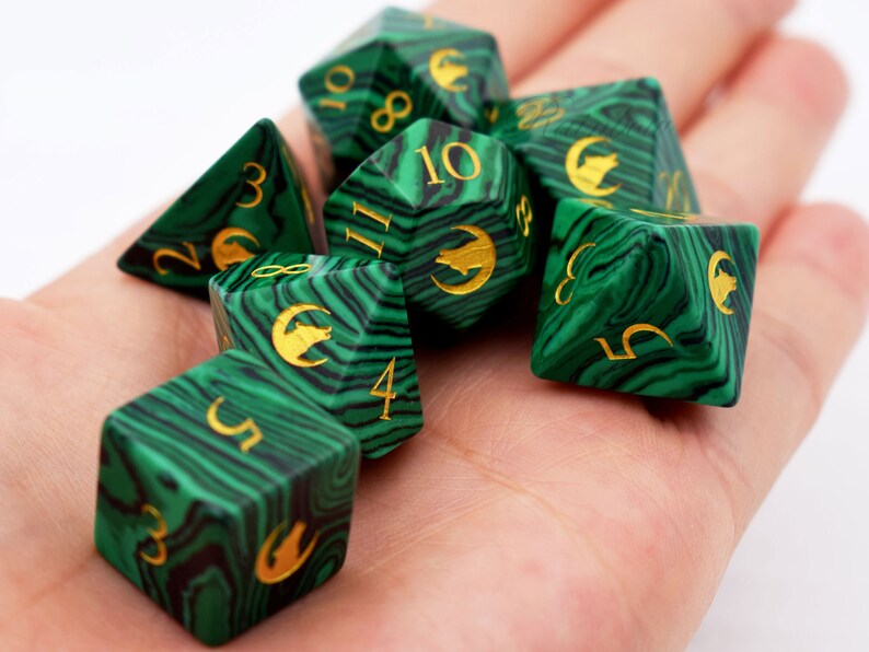 Puede incluir: Un conjunto de siete dados poli&eacute;dricos verdes y negros veteados, con n&uacute;meros dorados y s&iacute;mbolos de lobo. Los dados est&aacute;n dise&ntilde;ados para juegos de rol de mesa. Los dados se sostienen en una mano.