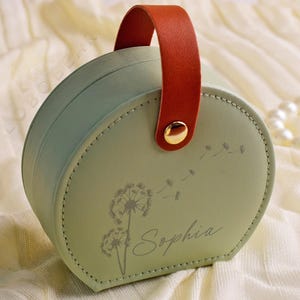 K&ouml;nnte beinhalten: Eine runde, mintgr&uuml;ne Schmuckschatulle mit braunem Griff und goldenem Knopfverschluss. Die Schatulle zeigt ein Pusteblumen-Design und den Namen "Sophia" in Schreibschrift. Sie liegt auf einer cremefarbenen Oberfl&auml;che.