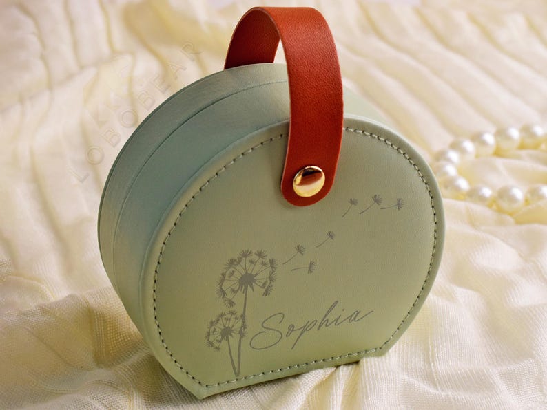 K&ouml;nnte beinhalten: Eine runde, mintgr&uuml;ne Schmuckschatulle mit braunem Griff und goldenem Knopfverschluss. Die Schatulle zeigt ein Pusteblumen-Design und den Namen "Sophia" in Schreibschrift. Sie liegt auf einer cremefarbenen Oberfl&auml;che.