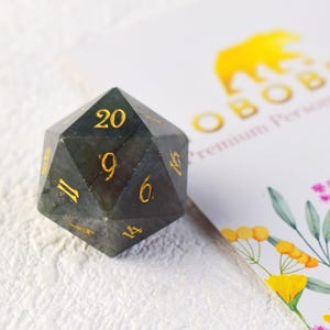 May include: A dark grey, 20-sided dice with gold numbers. The dice is made of a natural stone with a unique, iridescent pattern.