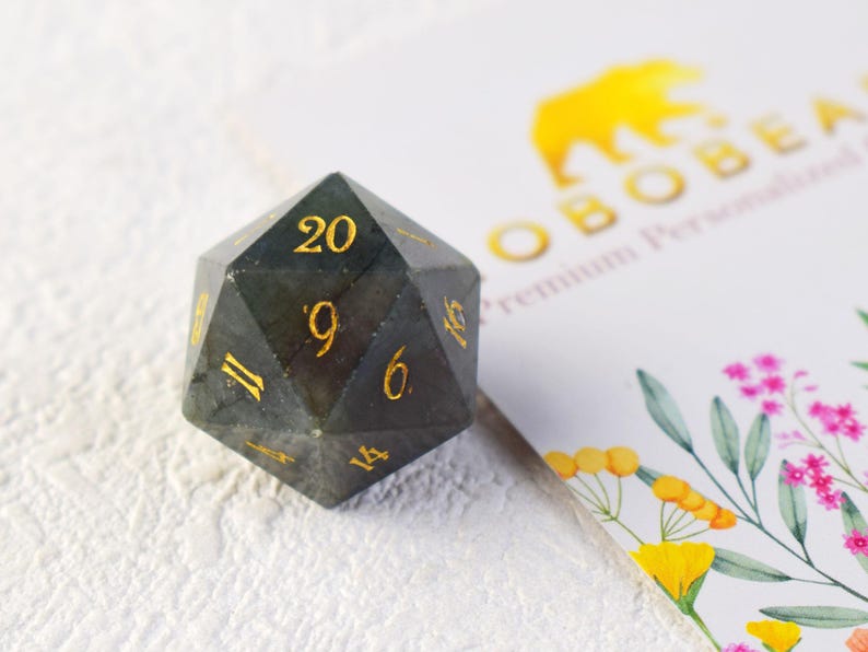 May include: A dark grey, 20-sided dice with gold numbers. The dice is made of a natural stone with a unique, iridescent pattern.