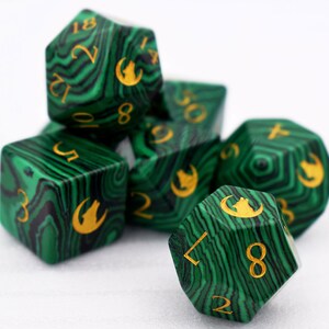 Puede incluir: Un juego de siete dados poli&eacute;dricos con un dise&ntilde;o verde y negro que imita la malaquita. Los dados tienen n&uacute;meros dorados y un s&iacute;mbolo de lobo. Apropiados para juegos de rol, miden unos 2-3 cm.