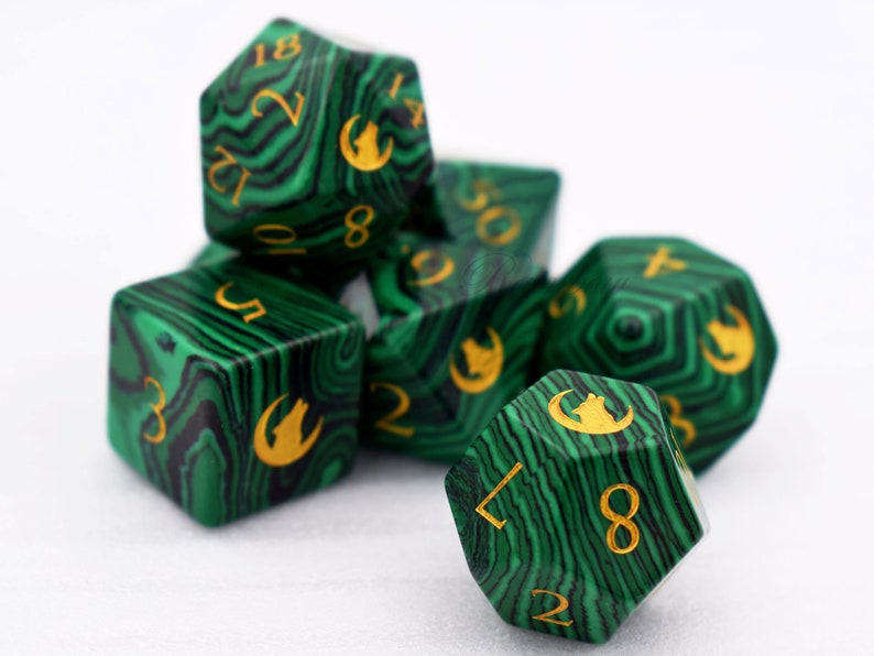 Puede incluir: Un juego de siete dados poli&eacute;dricos con un dise&ntilde;o verde y negro que imita la malaquita. Los dados tienen n&uacute;meros dorados y un s&iacute;mbolo de lobo. Apropiados para juegos de rol, miden unos 2-3 cm.