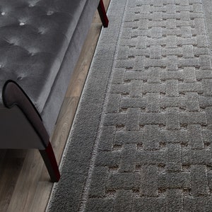 May include: A gray rectangular rug with a woven pattern, placed next to a gray upholstered bench. The rug has a textured surface and a border. The bench has button tufting and dark wood legs.