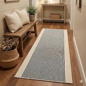 May include: A rectangular rug with a cream-colored border and black vertical stripes. The rug is placed on a wooden floor in a hallway, alongside a wooden bench with pillows and a woven basket.