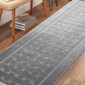 May include: A rectangular gray rug with a textured, woven pattern. The rug has a border and is placed on a hardwood floor. The rug's design features a repeating brick-like pattern.