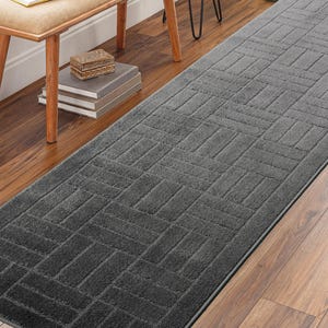 May include: A rectangular, dark gray rug with a geometric pattern. The rug is placed on a hardwood floor, and a bench and side table are visible in the background. The rug's design features a repeating rectangular block pattern.