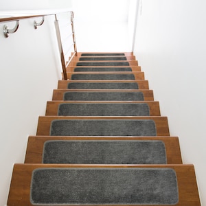 May include: A set of gray carpet stair treads with a non-slip surface. The treads are installed on wooden stairs with a natural wood finish.