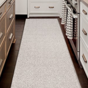 May include: A white kitchen runner rug with a textured surface. The rug is placed between a set of wooden cabinets and a set of white cabinets.
