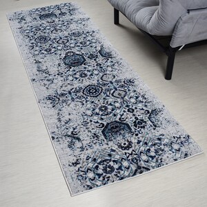 May include: A rectangular area rug with a distressed, ornate pattern in shades of gray, navy, and teal. The rug features a central design with floral and geometric motifs. The rug is placed on a light-colored wooden floor, with a gray sofa visible in the background.