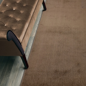 May include: A brown tufted bench with black legs sits on a wood floor in front of a brown area rug.