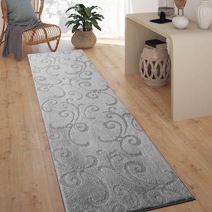May include: A rectangular, light gray runner rug with a scrolling, floral pattern. The rug is placed on a hardwood floor, and the design is a lighter shade of gray than the background. The rug is a decorative element for a hallway or entryway.