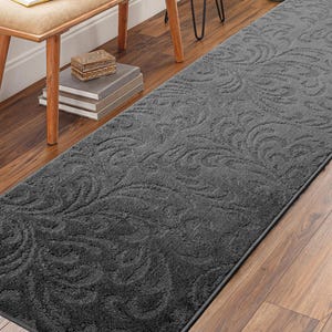 May include: A charcoal gray runner rug with a repeating floral pattern. The rug is rectangular and placed on a hardwood floor. The rug's design features a textured, raised pattern.
