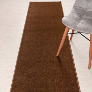 May include: A brown runner rug with a subtle texture. The rug is shown on a light wood floor with a chair in the background.