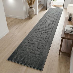 May include: A long, rectangular runner rug in a dark charcoal gray color. The rug features a repeating woven basket-weave pattern throughout the center, bordered by a solid, smooth-textured frame along all four edges of the floor covering.