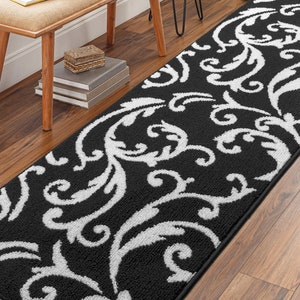 May include: Black and white patterned runner rug with a scrolling floral design. The rug is rectangular and placed on a hardwood floor. The runner has a black background with white floral patterns. The rug is near a wooden bench.