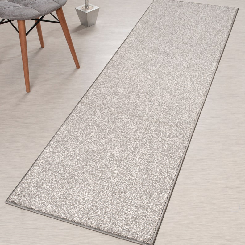 May include: A light gray runner rug with a subtle speckled pattern and a white border. The rug is laid out on a light wood floor.