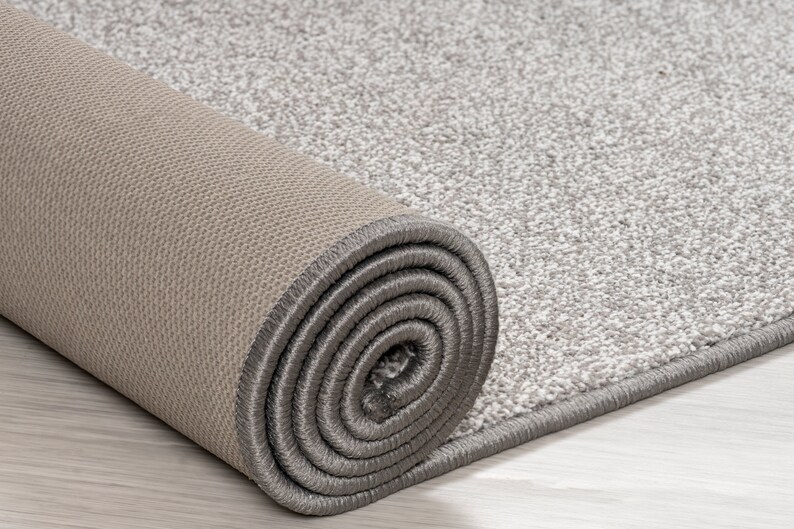May include: A close-up of a rolled-up gray and beige rug with a gray border. The rug is made of a soft, plush material.