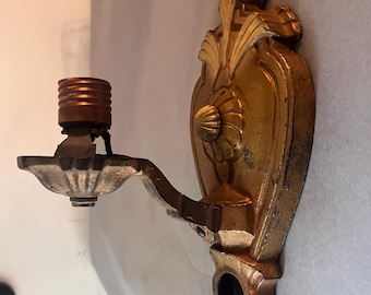 May include: Ornate gold-colored wall sconce with a decorative floral design. The sconce features a curved arm holding a light socket. The backplate has a detailed, textured surface. The fixture is designed for a single light bulb.