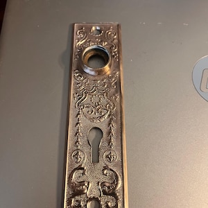 May include: Ornate antique bronze door plate with a keyhole and decorative floral and scrollwork. The rectangular plate features a circular opening and two stylized animal figures at the bottom. The plate is likely made of metal.