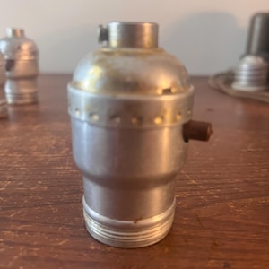 May include: A silver-colored lamp socket with a brown on/off switch. The socket has a cylindrical body with a threaded base and a small metal clip at the top. Other sockets are visible in the background.