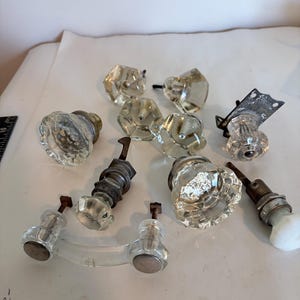 May include: A collection of vintage hardware including several clear glass doorknobs with faceted surfaces, a glass drawer pull with metal ends, and various metal mounting components and spindles, some showing signs of oxidation and age.