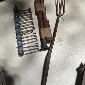 May include: An assortment of antique tools, including a metal hay fork, a rack of metal hooks with blue and gray accents, and two wooden rods bound with tan fabric.