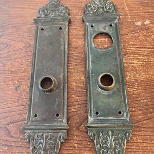 May include: Two antique brass door plates with ornate designs. The plates have a rectangular shape with a rounded top and bottom. The plates have a keyhole and a knob hole.