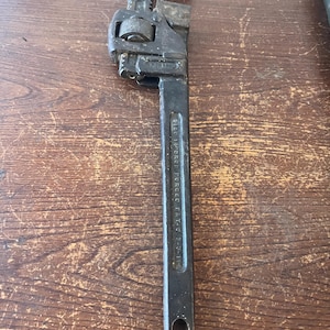 May include: A black pipe wrench with a textured jaw and adjustable mechanism. The wrench is made of metal and has a long handle with a hole at the end. The text "SIZE 14 PIPE" is visible on the handle.