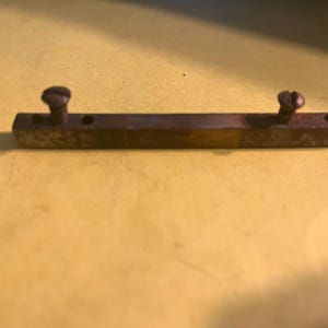 May include: A rectangular metal bar with two screws and two holes. The bar is a dark, aged brown color, with the screws appearing to be the same color. The background is a light yellow.