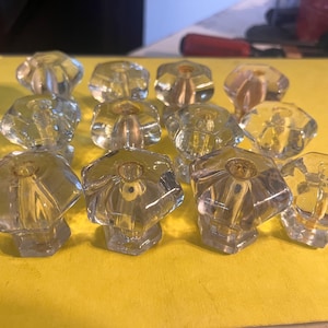 May include: A collection of twelve clear glass cabinet knobs with a faceted, flower-like design. The knobs are arranged on a yellow surface, showcasing their transparent quality and decorative shape. These knobs are suitable for furniture restoration or DIY projects.