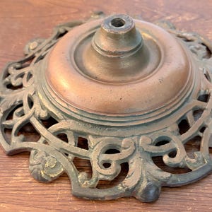 May include: An antique bronze-colored metal lamp base with an ornate, openwork design around the perimeter. The base has a circular top with a central hole for a lamp post. The metal has a patina.