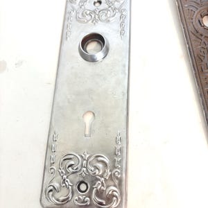 May include: A rectangular silver-toned metal door plate featuring embossed scrollwork patterns at the top and bottom, a central circular opening for a knob, and a lower keyhole cutout. The plate includes two small screw holes for mounting purposes.