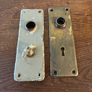 May include: Two antique door plates, one is brass and the other is painted white. Both have a keyhole and a round hole for a knob.