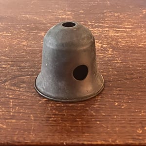 May include: A small, bell-shaped metal lampshade with a dark, aged finish. It features a circular opening at the top and a larger, round hole on the side. The lampshade is sitting on a wooden surface.