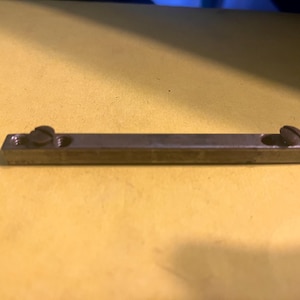 May include: A rectangular metal object with two screws. The object is a dark, dull gray color and appears to be made of metal. The screws are also dark and appear to be made of metal. The object is lying on a yellow surface.