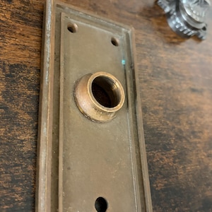 May include: A vintage bronze door plate with a keyhole and a circular opening for a doorknob. The rectangular plate has a raised border and two screw holes. The metal shows signs of age and wear.