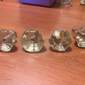 May include: Four clear, faceted glass cabinet knobs with metal hardware. The knobs have a hexagonal shape and are arranged on a wooden surface. The knobs are vintage and have a classic design.